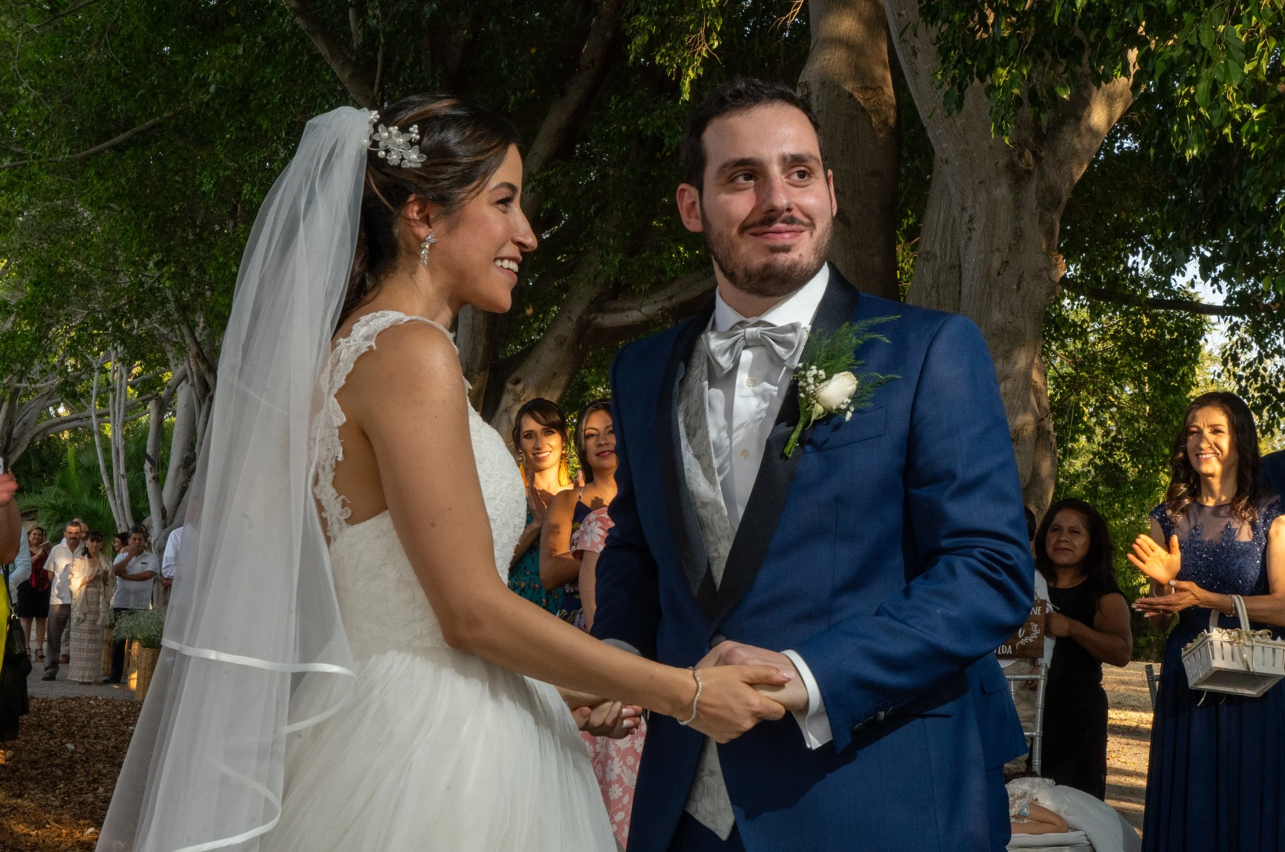 Una pareja de recién casados en su boda, con la novia usando un vestido blanco y el novio con un esmoquin azul, rodeados de familiares y amigos en un entorno al aire libre con árboles.