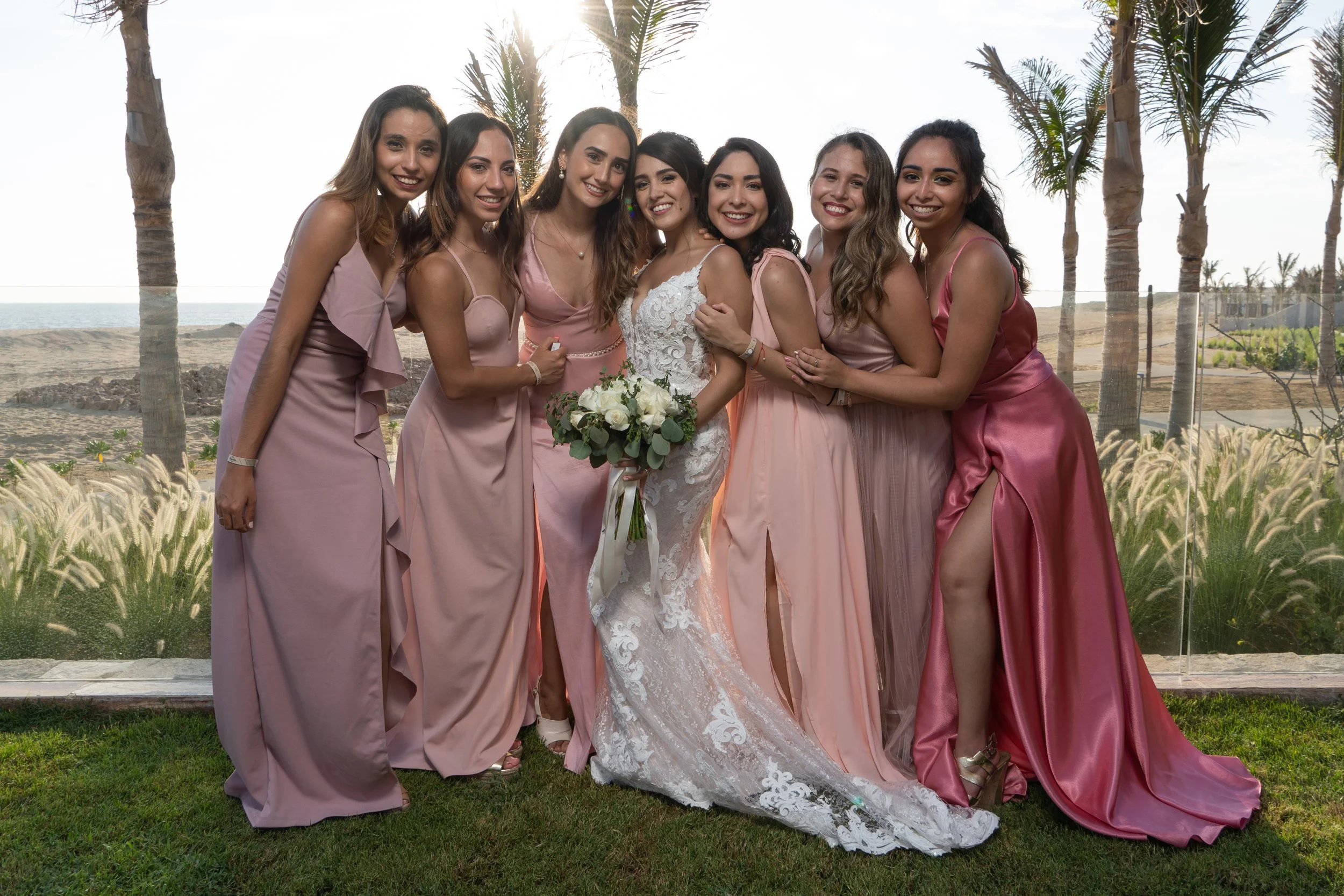 Grupo de ocho mujeres en un escenario al aire libre, con fondo de palmeras, playa y mar, vestidas con vestidos de diferentes tonos rosados, posando y sonriendo para una foto, con una de ellas en el centro con vestido de novia y ramo de flores.