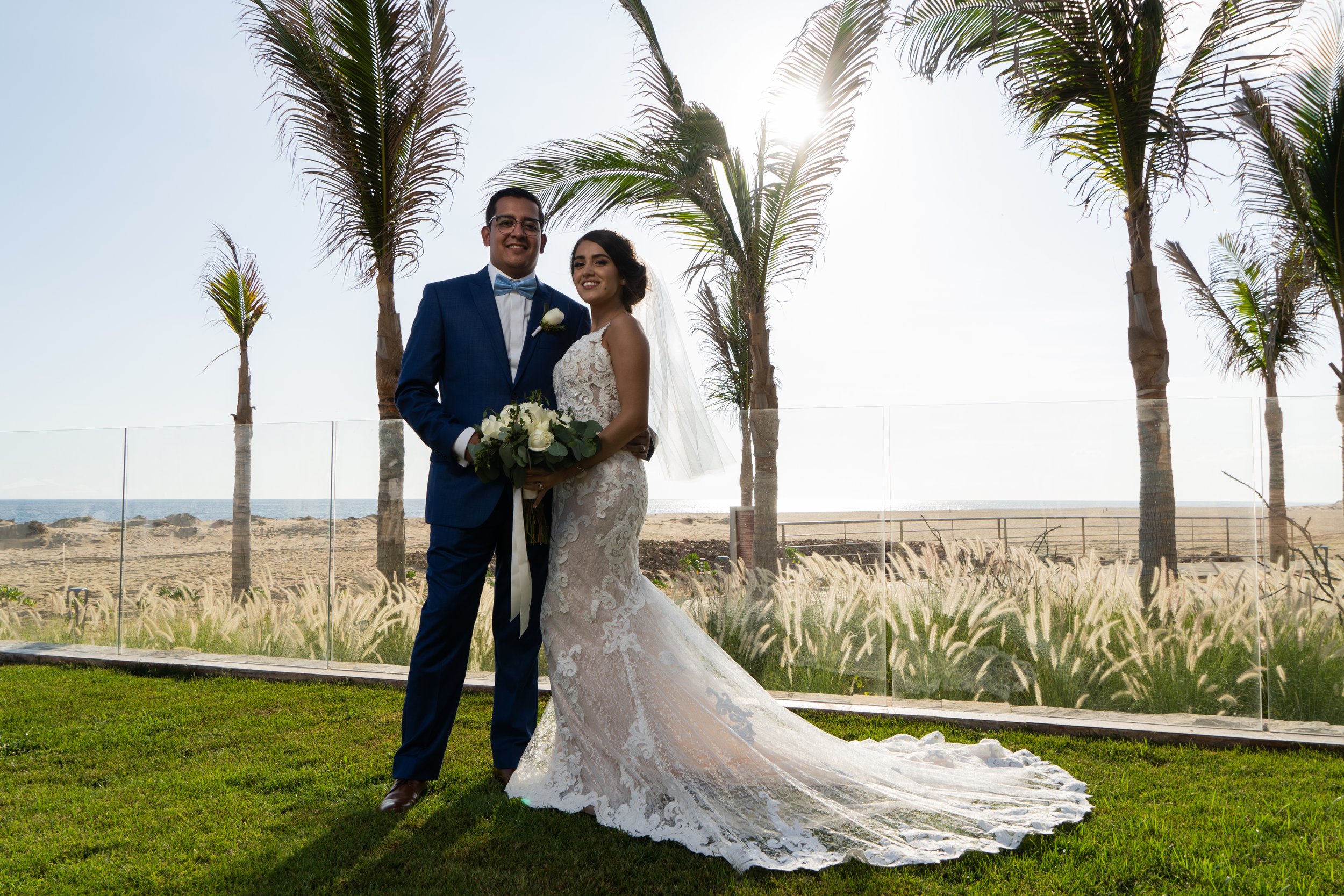 Una pareja de recién casados en la playa con palmeras, la novia con vestido de encaje y el novio con traje azul y moño, sosteniendo un ramo de flores blancas.