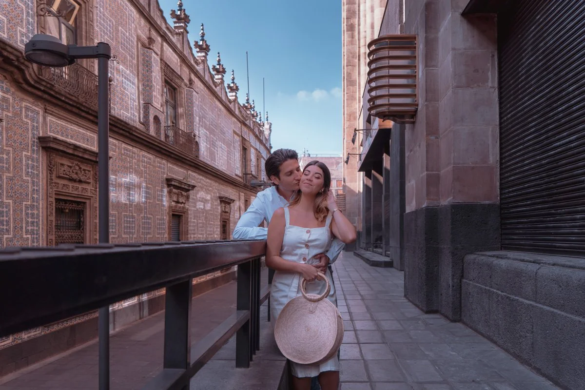 A young couple standing close together on a city sidewalk with historic and modern buildings behind them, the man kissing the woman on her temple, her eyes closed, she holding a straw handbag and touch her hair.