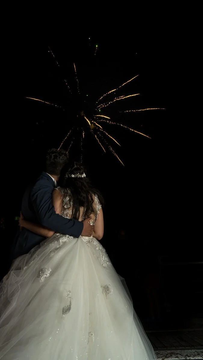 A bride and groom sharing a moment during their wedding celebration at night, with fireworks illuminating the sky.