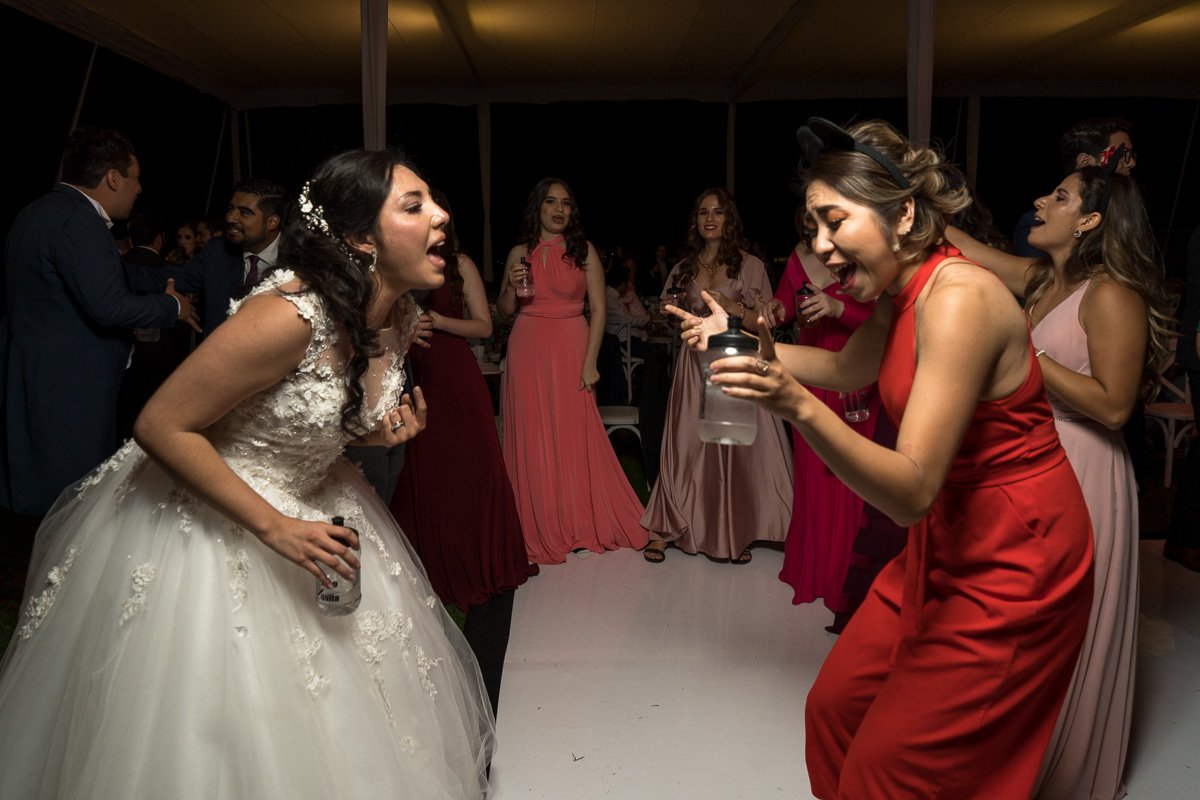 Wedding reception scene with women dancing and singing, including a bride in a white wedding dress and a woman in a red dress, at night indoors
