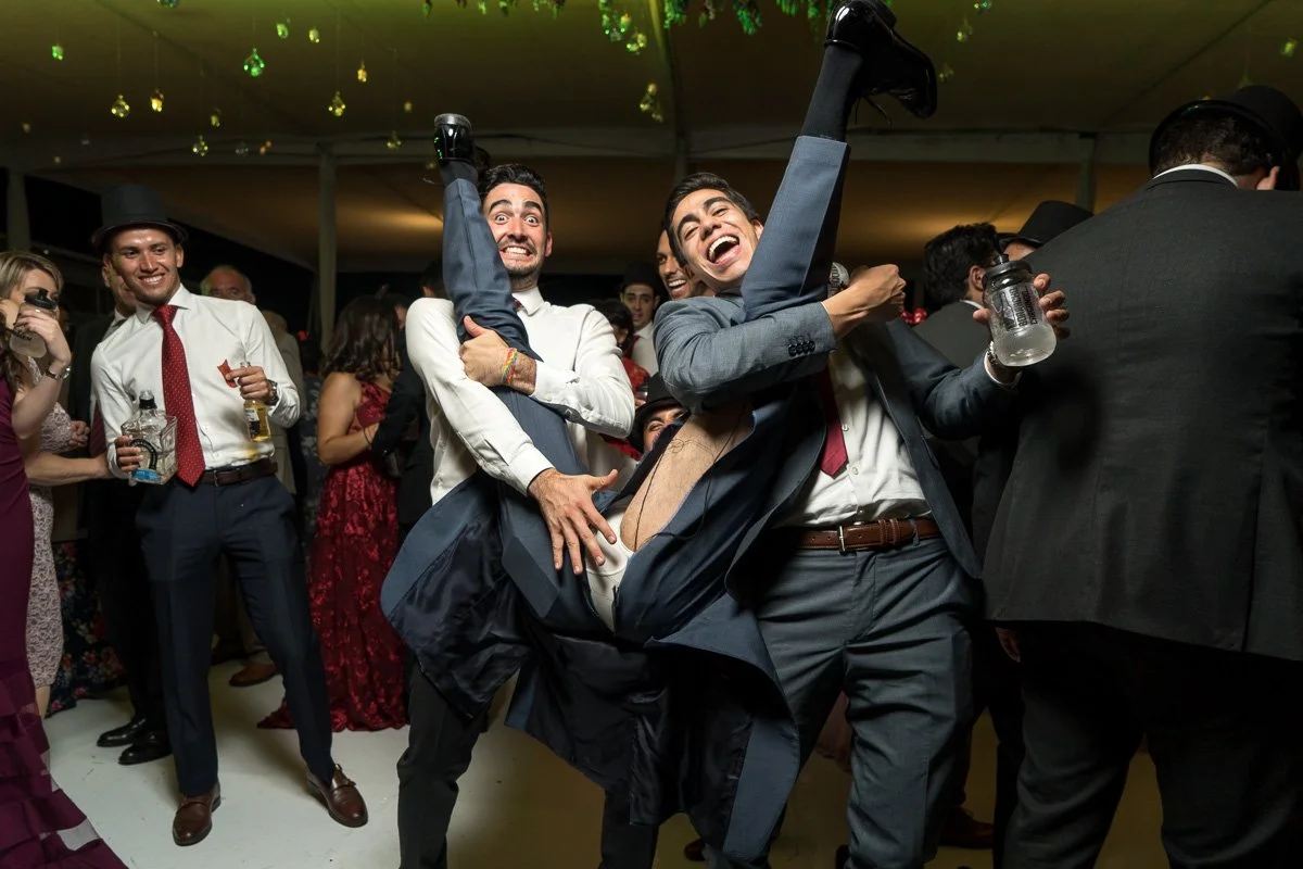 Group of people dancing and celebrating at a party or wedding reception, with two men lifting a third man in a joyful display of fun.