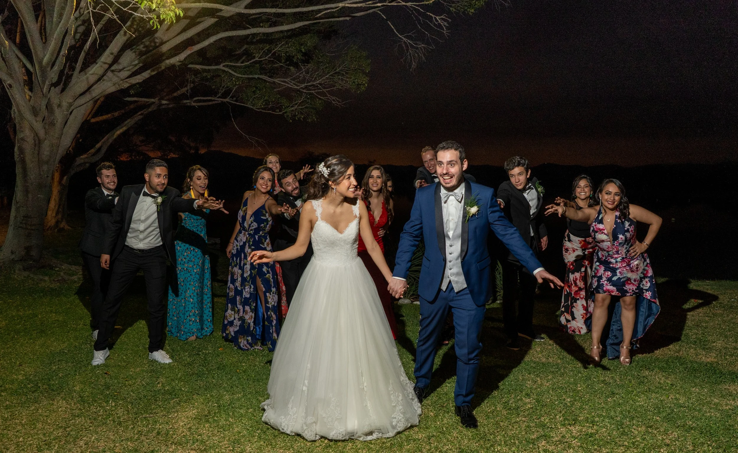 Grupo de personas en una boda al aire libre en la noche, sonriendo y bailando, con un árbol grande a la izquierda y un cielo oscuro.