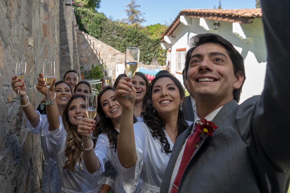 A wedding celebration with the bride and groom taking a selfie, surrounded by smiling guests holding champagne glasses outdoors near a stone wall and a house with a tiled roof.