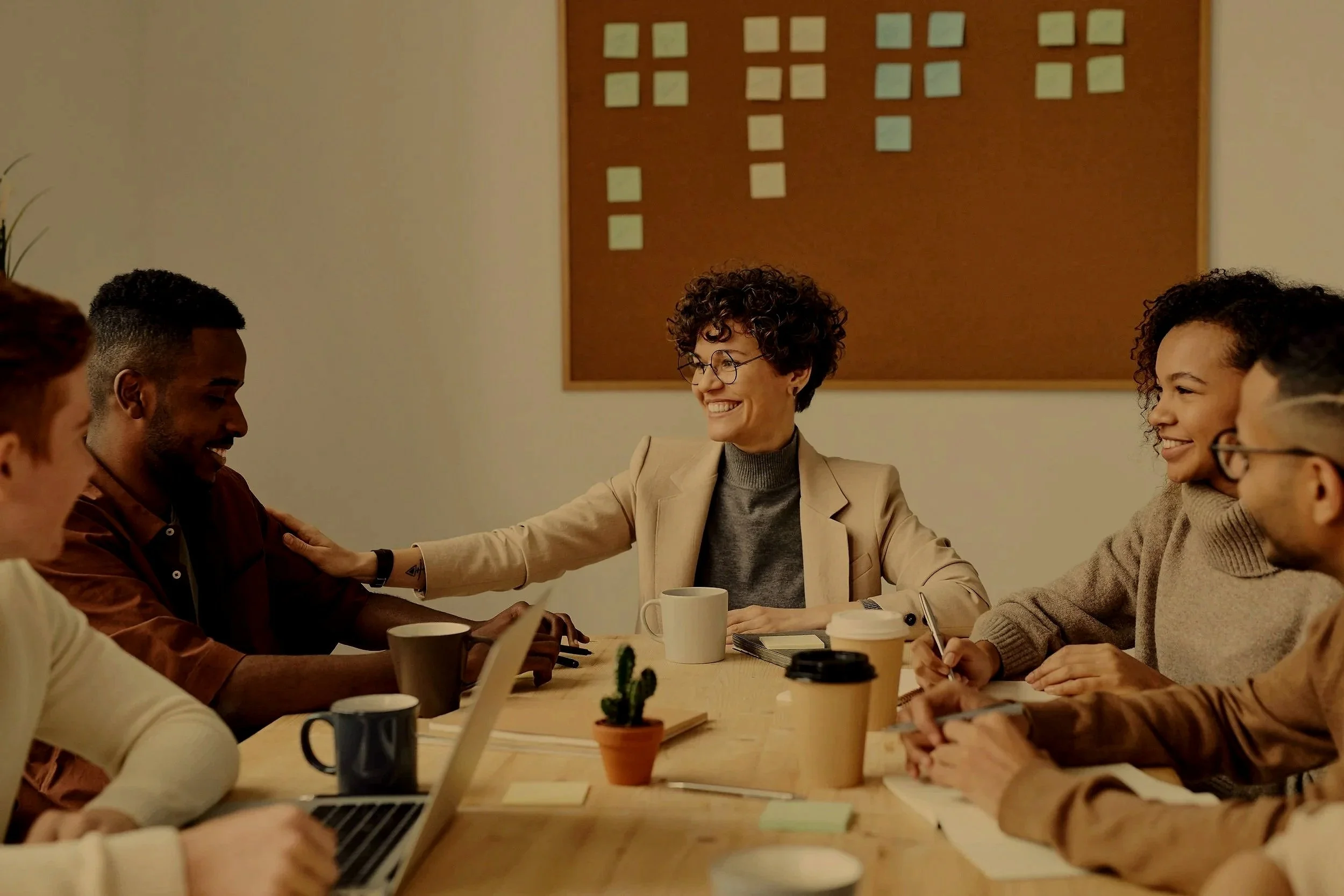 Group of diverse colleagues in a meeting room, smiling and engaging with each other around a wooden table, with laptops, coffee cups, and notebooks, in a professional office setting.