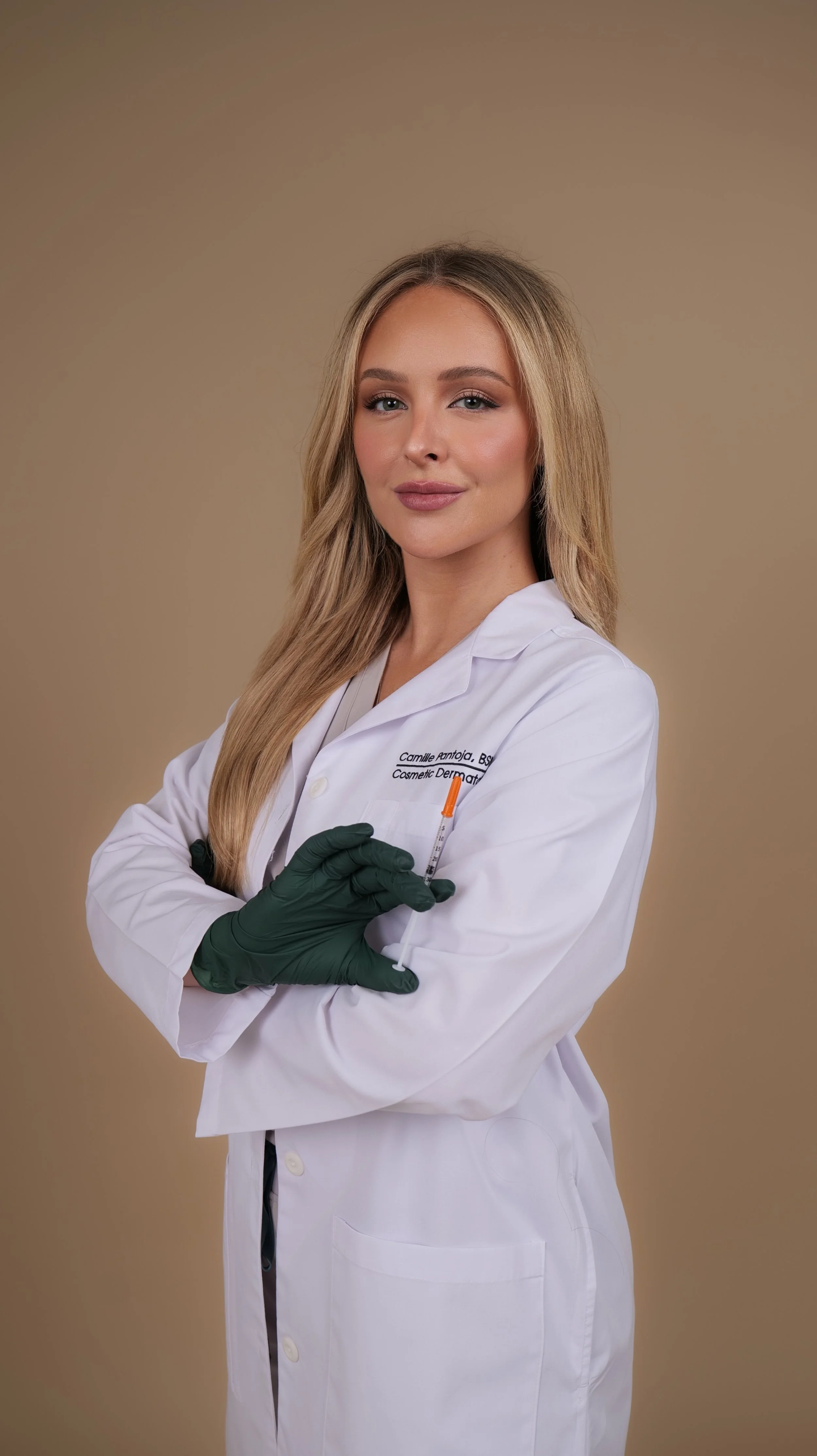 A woman in a white medical coat with her arms crossed, holding a syringe with orange liquid, wearing green gloves, standing against a neutral background.