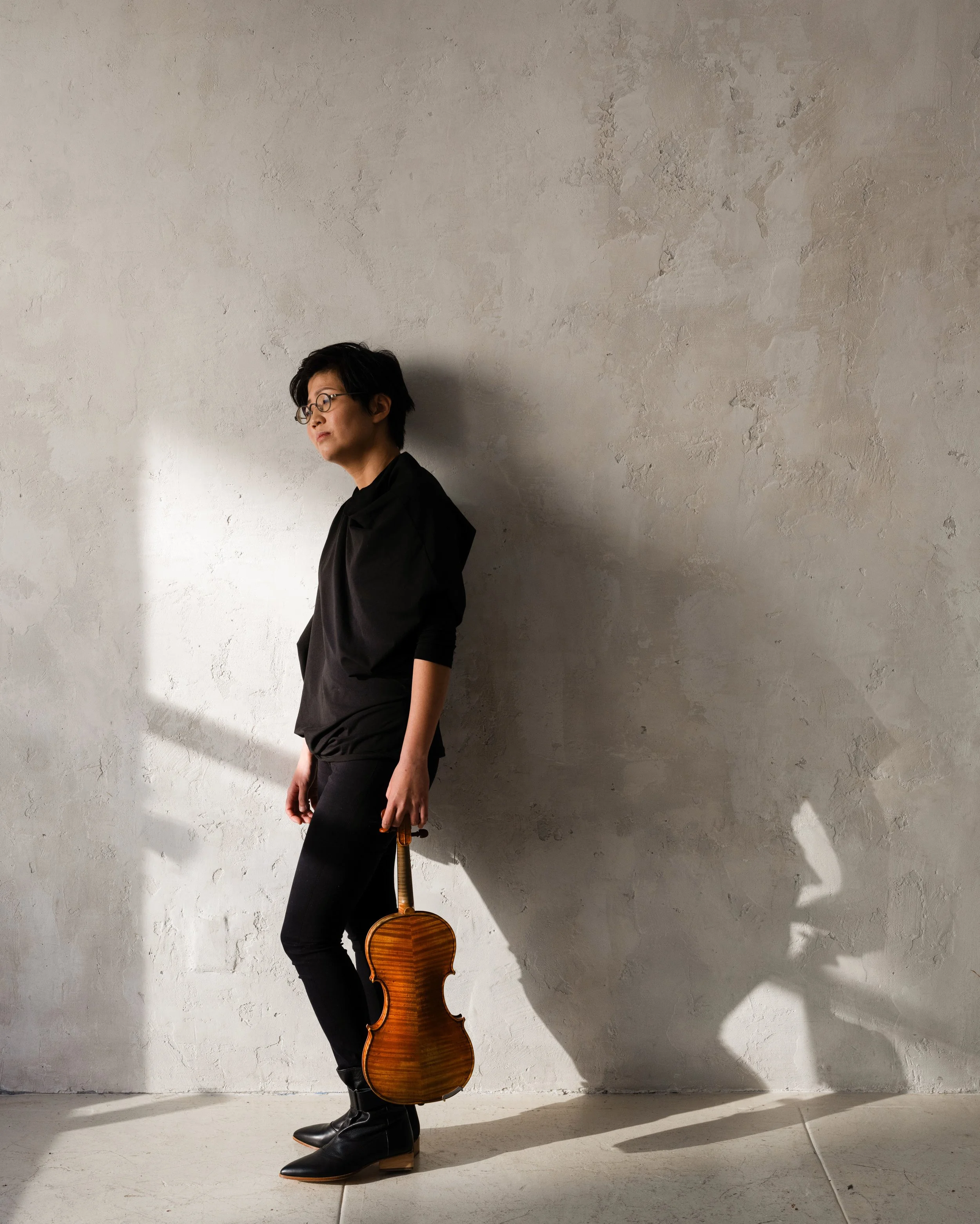 A woman with short black hair and glasses is standing against a textured light gray wall, holding a wooden violin by her side. She is wearing a black outfit and black boots, looking contemplative.