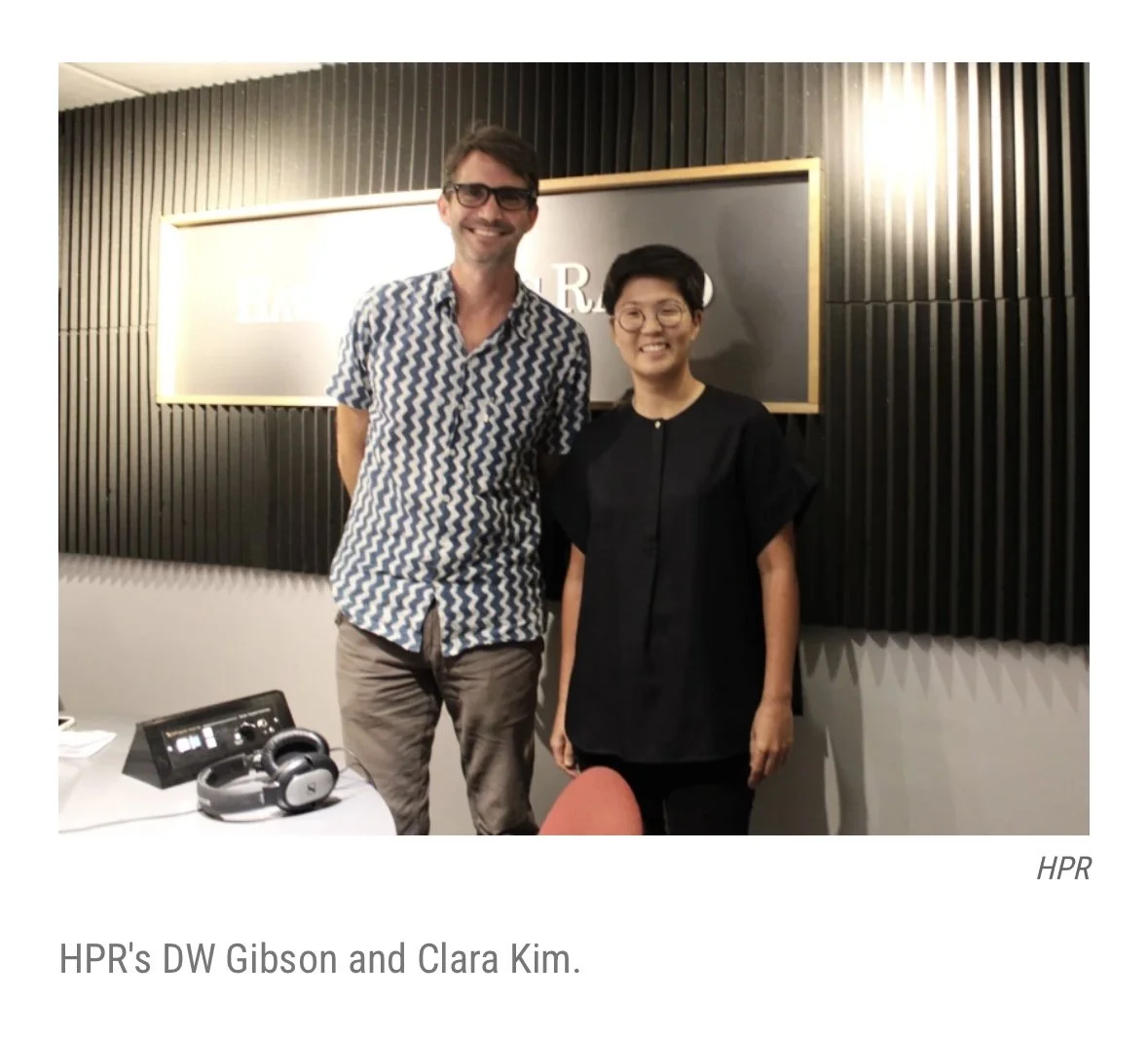 Two people, a man and a woman, standing side by side smiling in a room with a black and gold wall behind them. The man is wearing glasses and a patterned short-sleeve shirt, and the woman is wearing glasses and a black top. There is a table with headphones and electronic equipment in front of them. The caption reads: "HPR's DW Gibson and Clara Kim."
