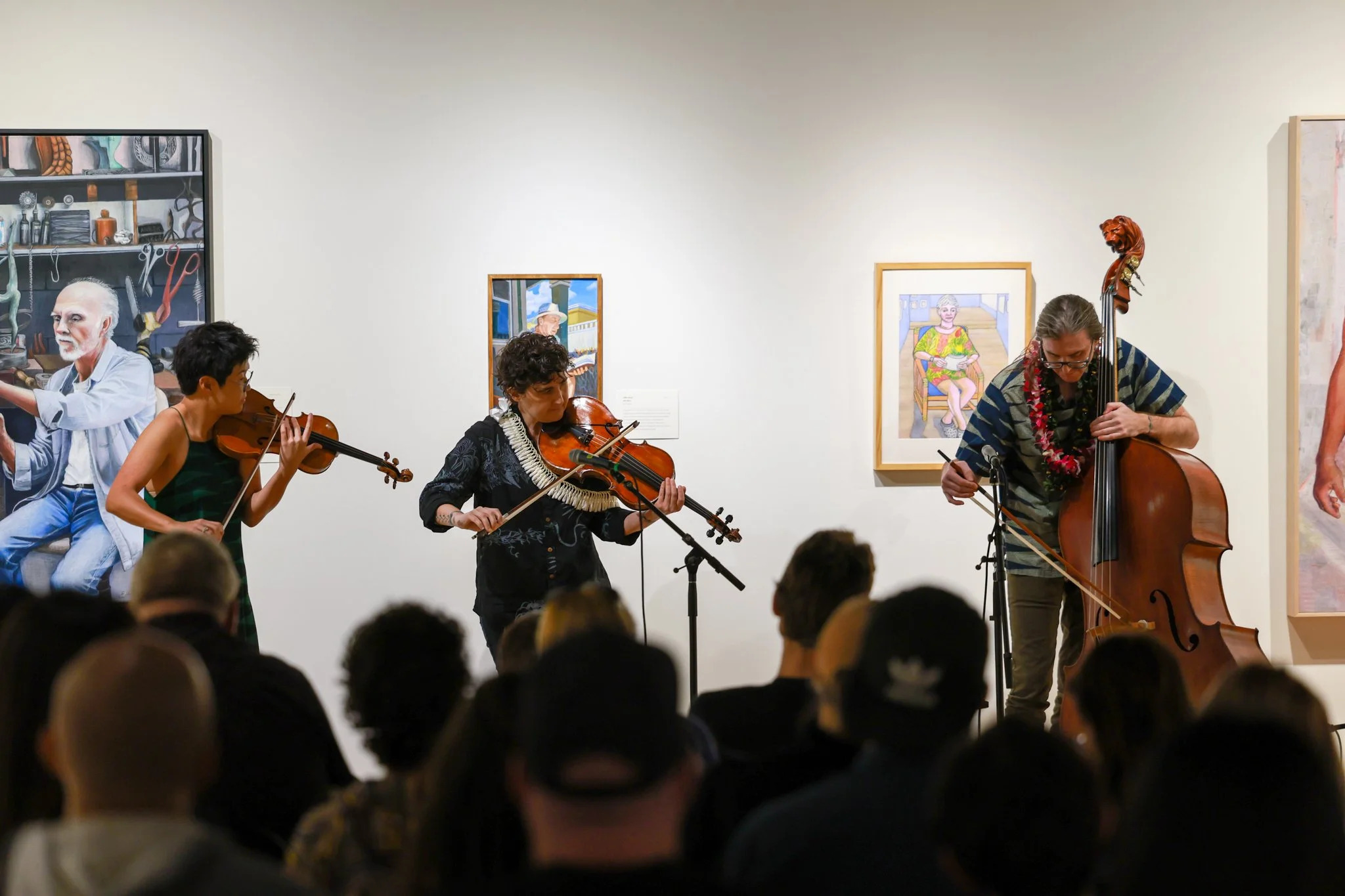 Three musicians performing with violins and a double bass in an art gallery, with paintings on the wall behind them and an audience watching.