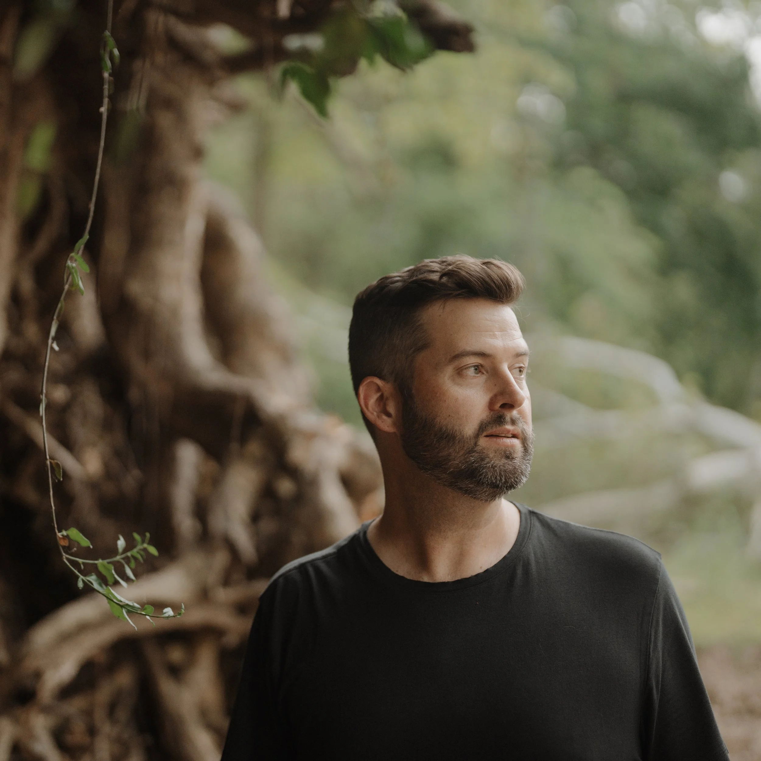 A man with a beard and short hair looking to the side, standing outdoors near a tree with green foliage.