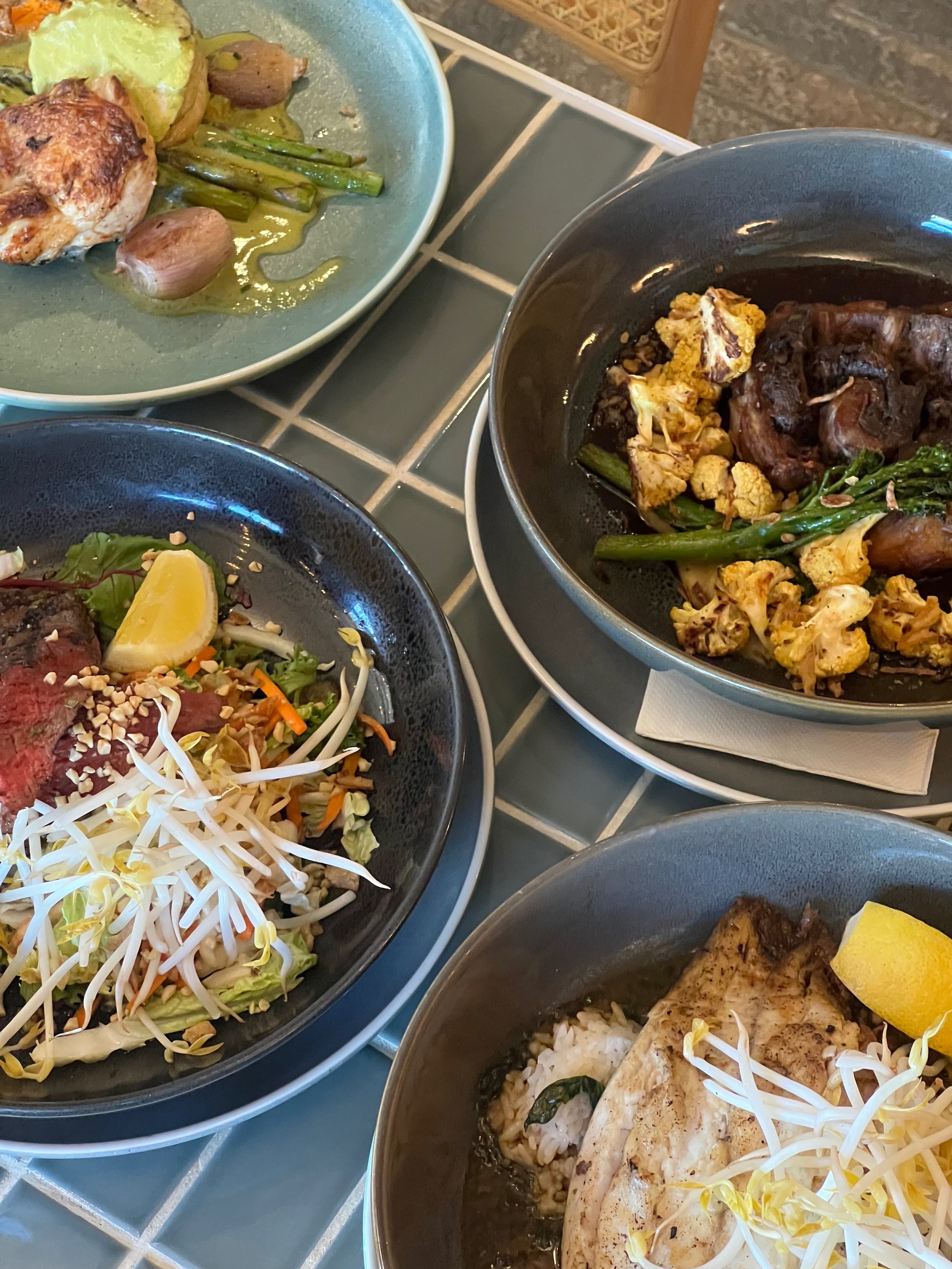 Assorted dishes including a Thai Kangaroo Salad, Lam Roulette, Barramundi and Chicken Breast on a green tiled table.