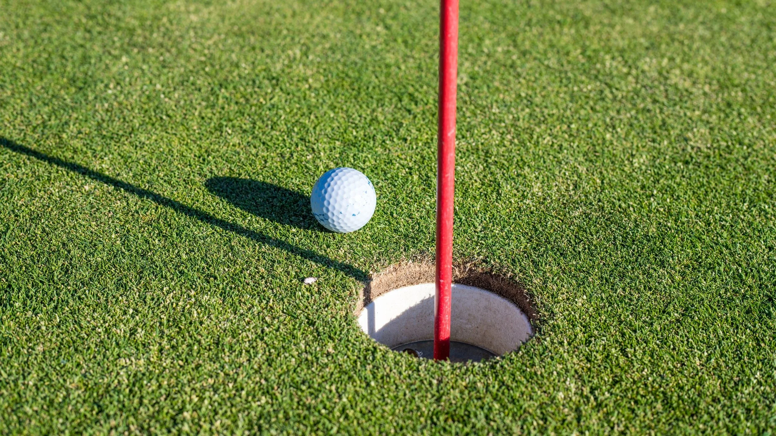 Close-up of a golf ball near the hole on a putting green, with a red flagstick marking the hole.