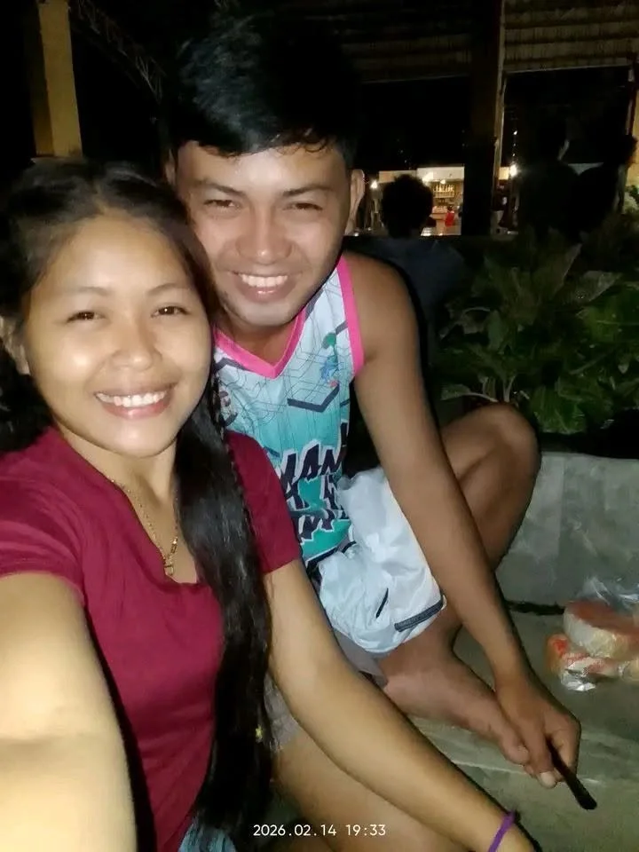 A young woman and a young man are taking a selfie together, smiling at the camera. The woman has long dark hair and is wearing a red shirt, while the man has short dark hair and is wearing a sleeveless shirt with colorful graphics. They are seated ou
