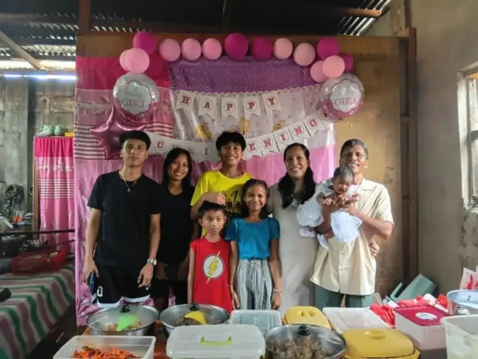 Family celebration with birthday decorations, pink and purple balloons, and a banner that says 'Happy Birthday' in a rustic room with seven people, including children and a baby, standing and smiling.