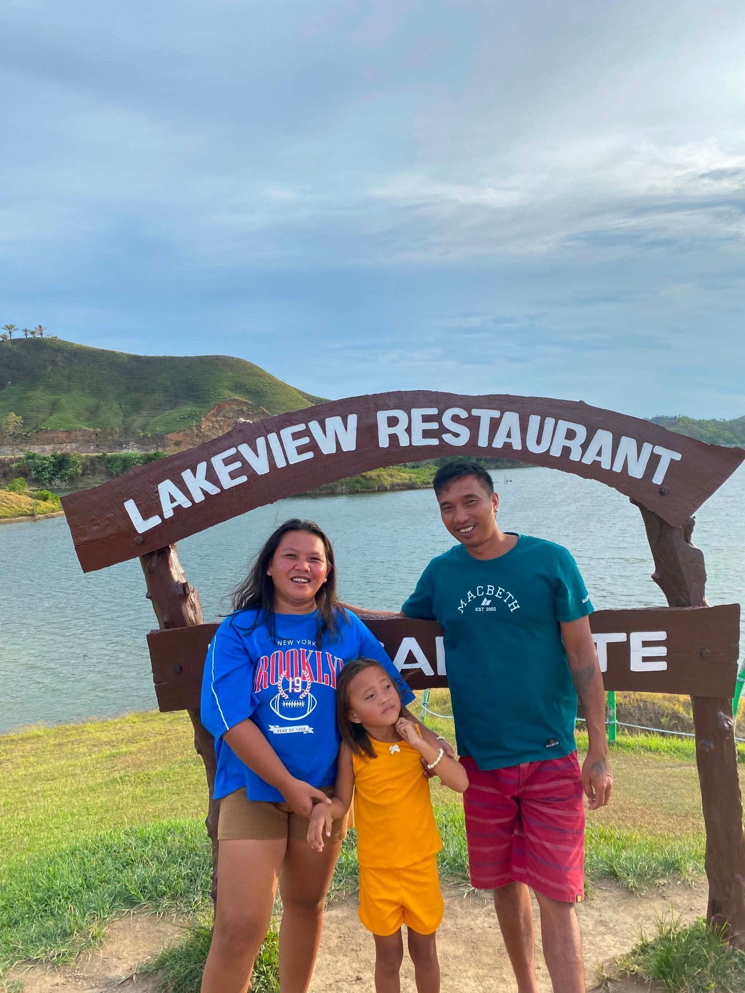 Family of four standing in front of a lake with green hills and a sign that reads 'Lakeview Restaurant' at Lake A Lake, smiling at the camera on a clear day.