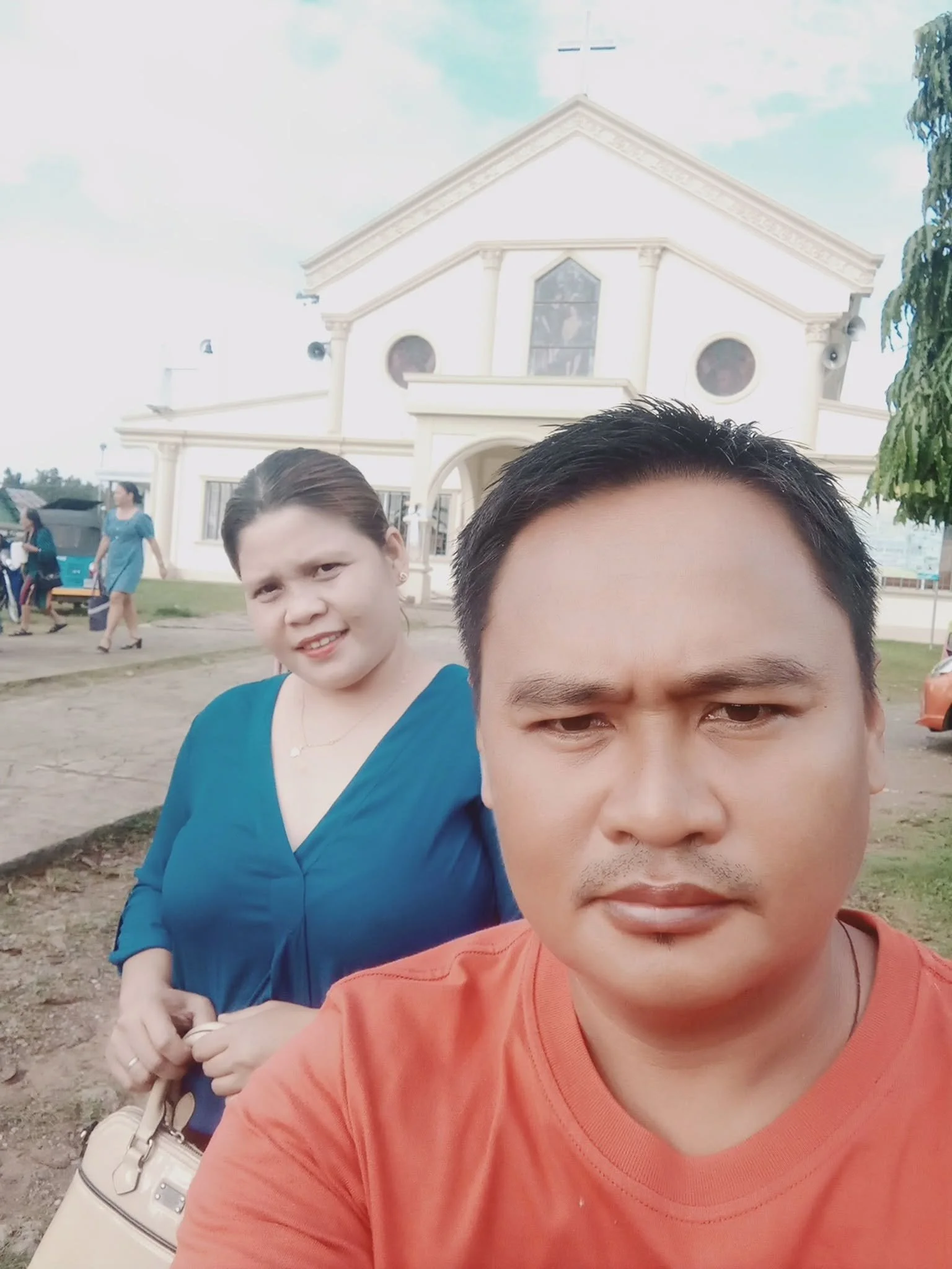Two people taking a selfie in front of a large white building with columns and a tower, with a woman holding a purse and other people in the background.