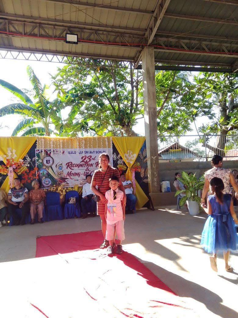 A woman and a young girl standing on a red carpet at a recognition event at a school. The background features a decorated banner, chairs with seated people, and lush trees, indicating a school auditorium or open-air space.