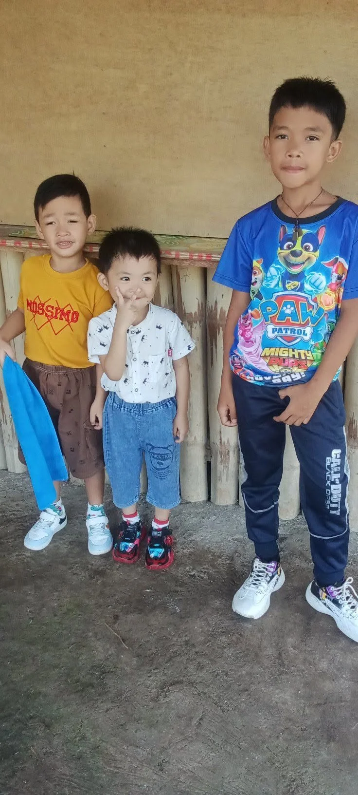Four boys standing outside against a simple wall and dirt ground. The youngest boy in the middle is making a peace sign. The boy on the right is wearing a blue Paw Patrol T-shirt. The boy on the left is wearing a yellow shirt and brown shorts. The yo