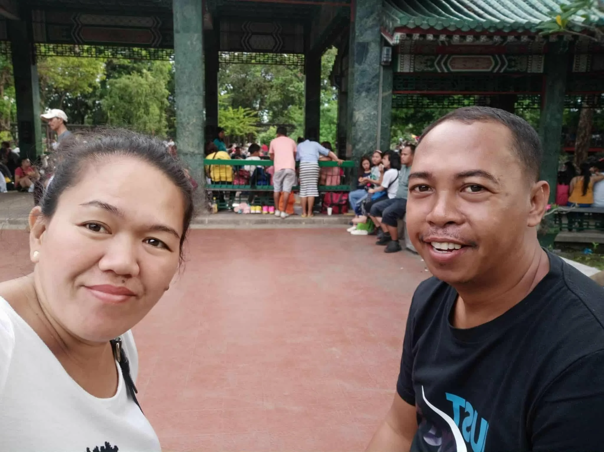 Close-up of a woman and a man taking a selfie at a park with traditional Asian architecture and many people in the background.