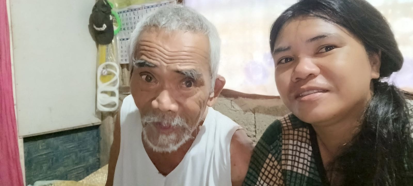 A close-up selfie of an elderly man with white hair and beard next to a woman with dark, wavy hair, inside a room with a patchwork wall and household items in the background.