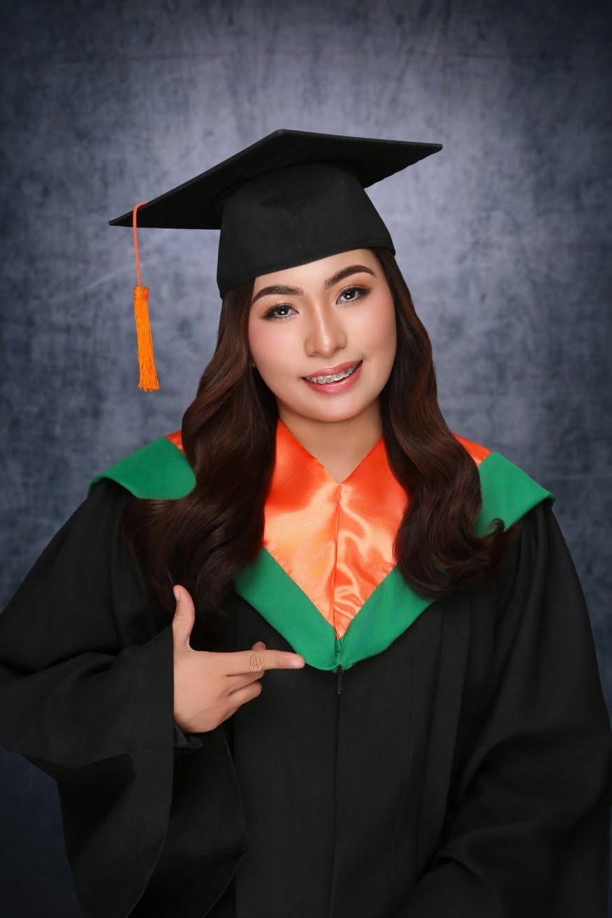 A young woman wearing a black graduation cap and gown, with an orange and green hood, smiling and pointing at her gown against a dark textured background.