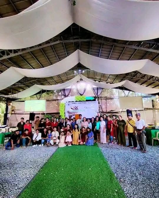 Group of people gathered at an indoor event under draped fabric ceiling, with a green carpet leading to a stage with screens and banners.