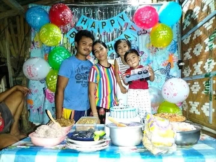 A family celebrating a birthday indoors with colorful balloons, a birthday cake, and a child holding a boombox toy. Four family members are posing behind a table with food and gifts, with a "Happy Birthday" banner in the background.