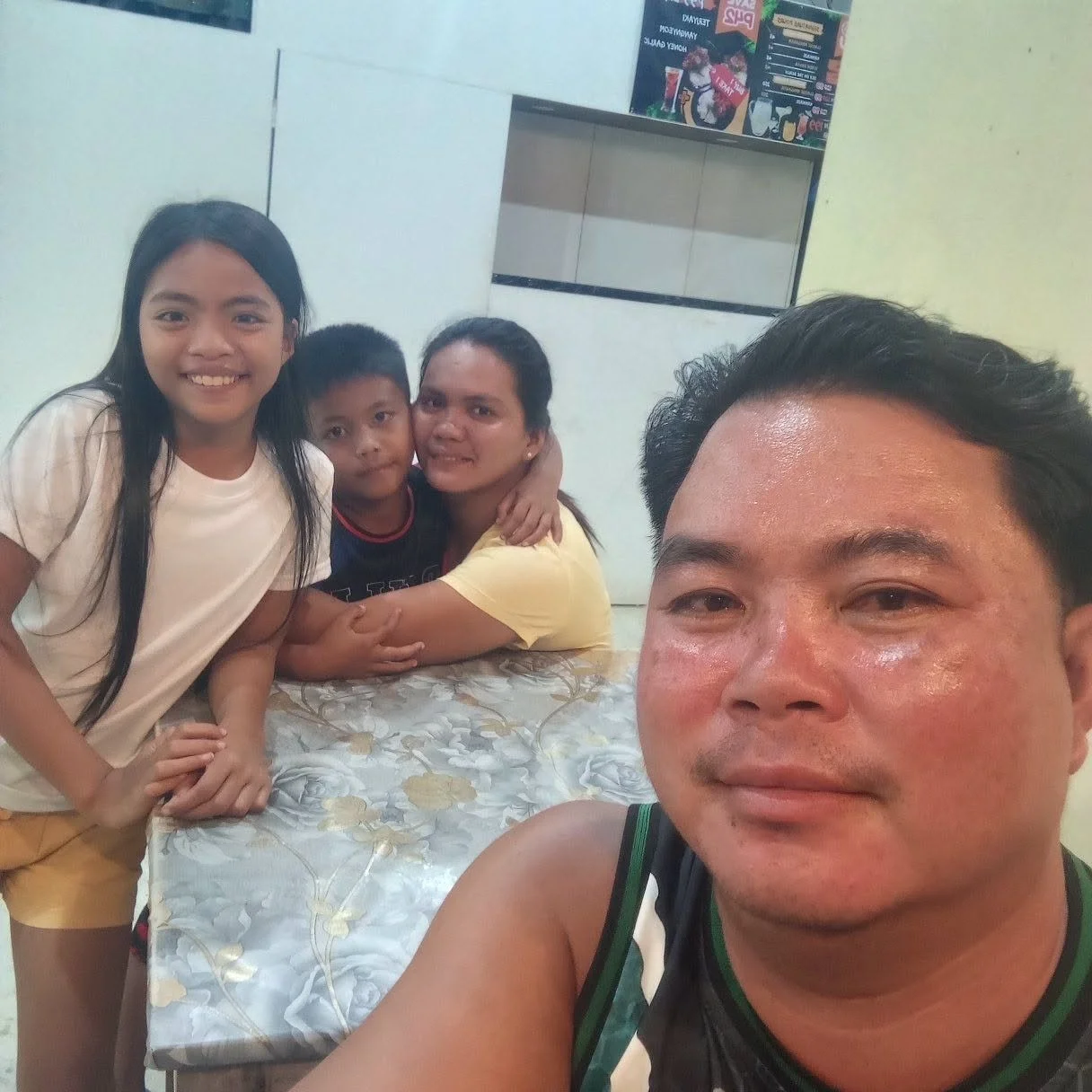 Four people, including a man taking a selfie, a woman, and two children, inside a room with a table covered in a floral tablecloth and a wall with a menu or advertisement in the background.