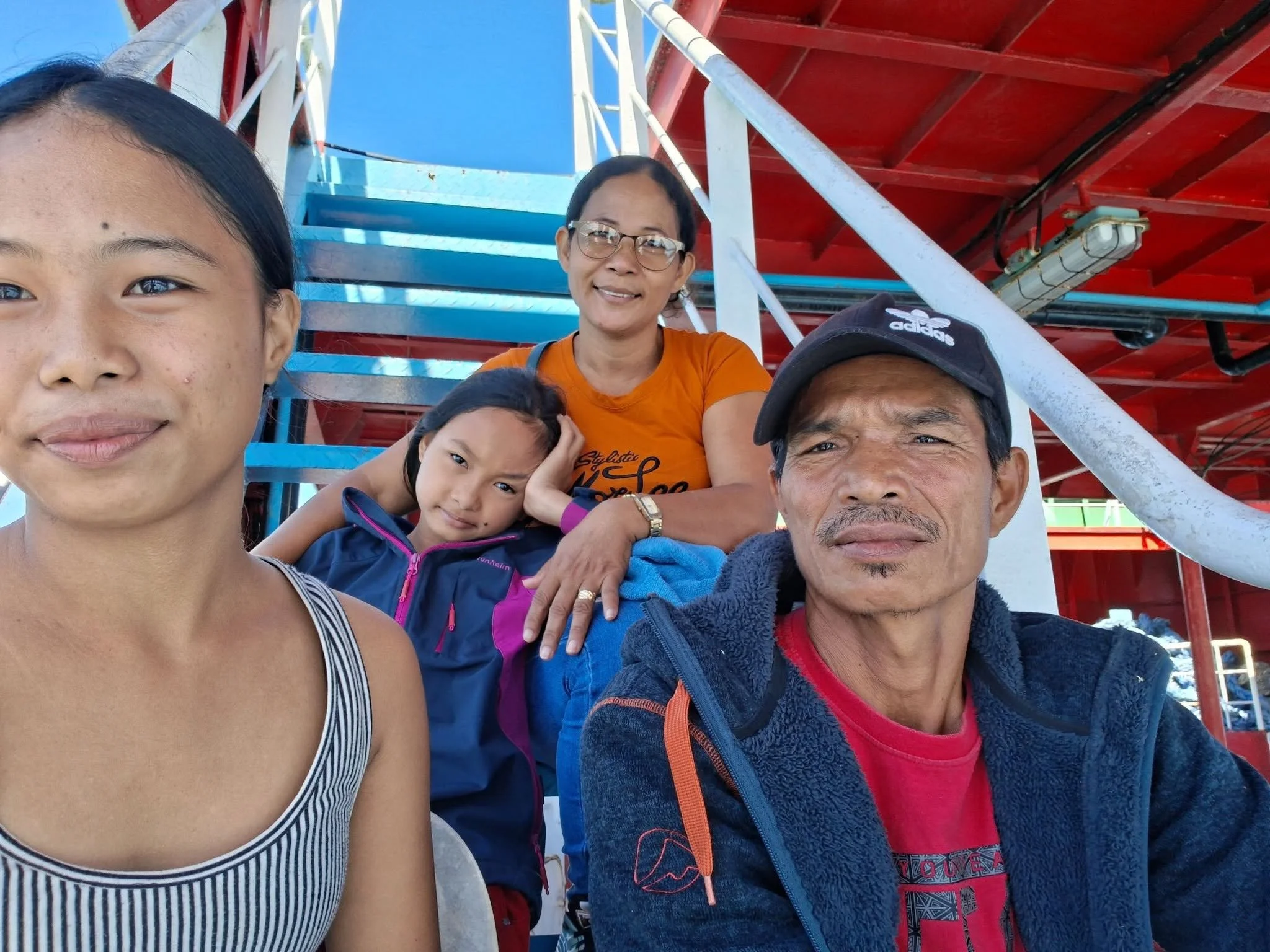 Group of five people, including a woman with glasses, a man with a cap, two young girls, and a young woman, taking a selfie on a Ferris wheel with red and blue structure and a clear blue sky