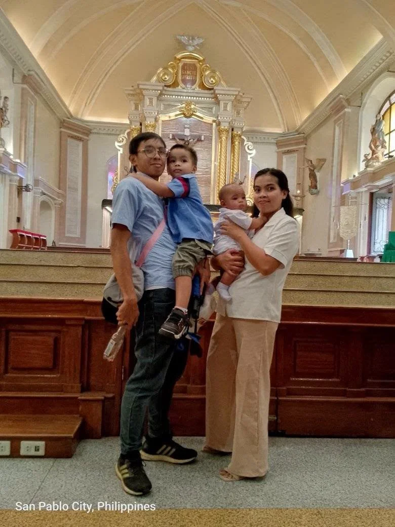 Two women and two children standing inside a church in San Pablo City, Philippines. The woman on the left is holding a young boy, and the woman on the right is holding a baby. The church has ornate gold and white decorations and high arched ceilings.
