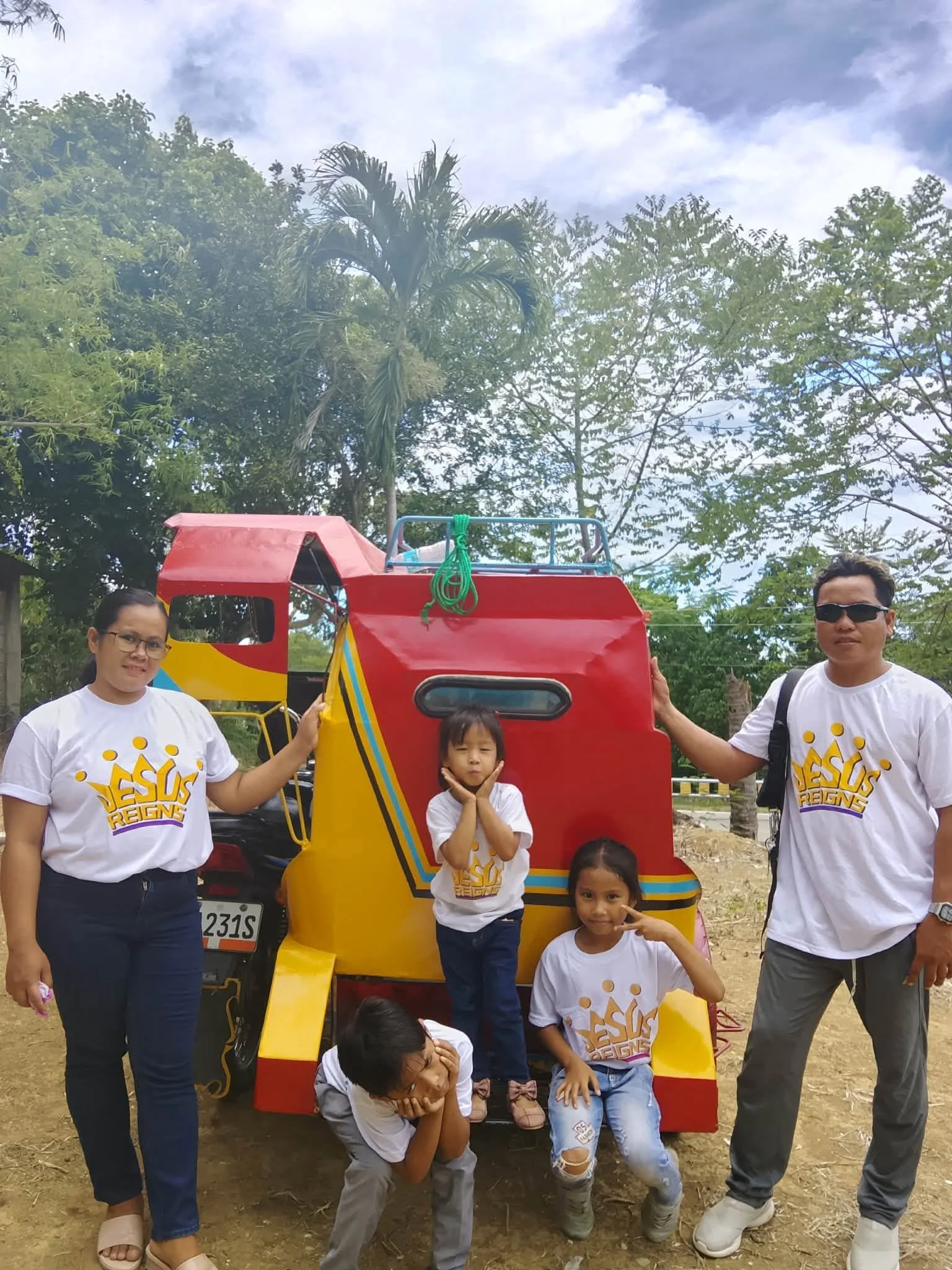 Family group standing outdoors in front of a colorful tricycle vehicle, with some children and adults smiling and posing for the photo, surrounded by trees and a cloudy sky.