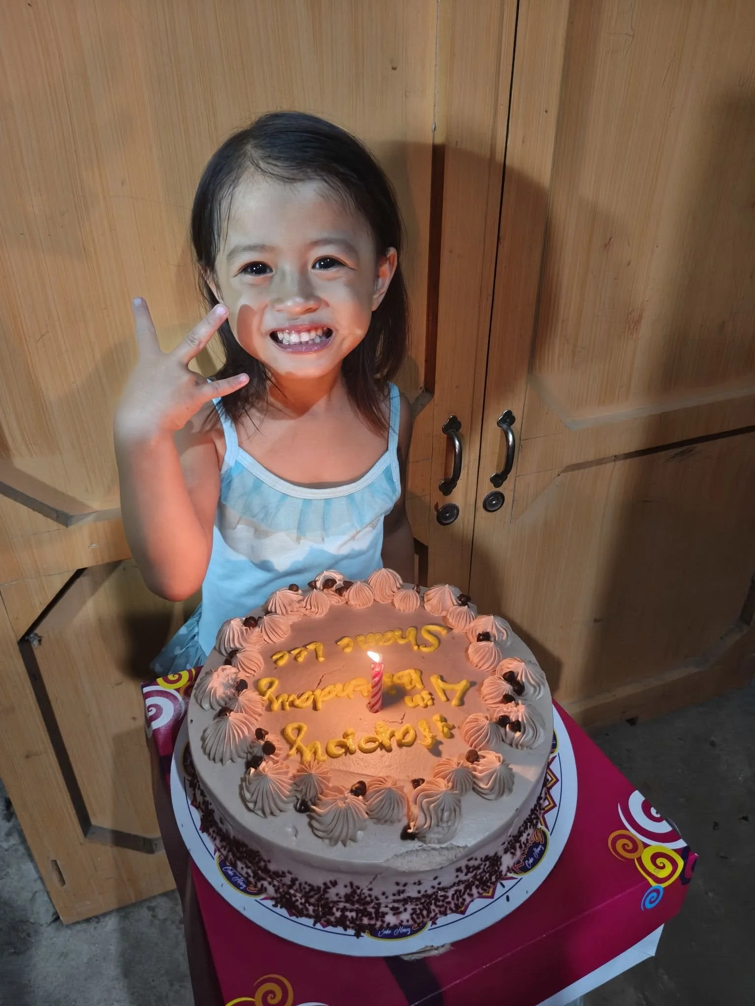 A young girl wearing a blue dress is celebrating her birthday, standing next to a large birthday cake with pink frosting and a lit birthday candle. The cake has yellow writing that says 'Happy Birthday Shaicha' and is decorated with purple and brown 