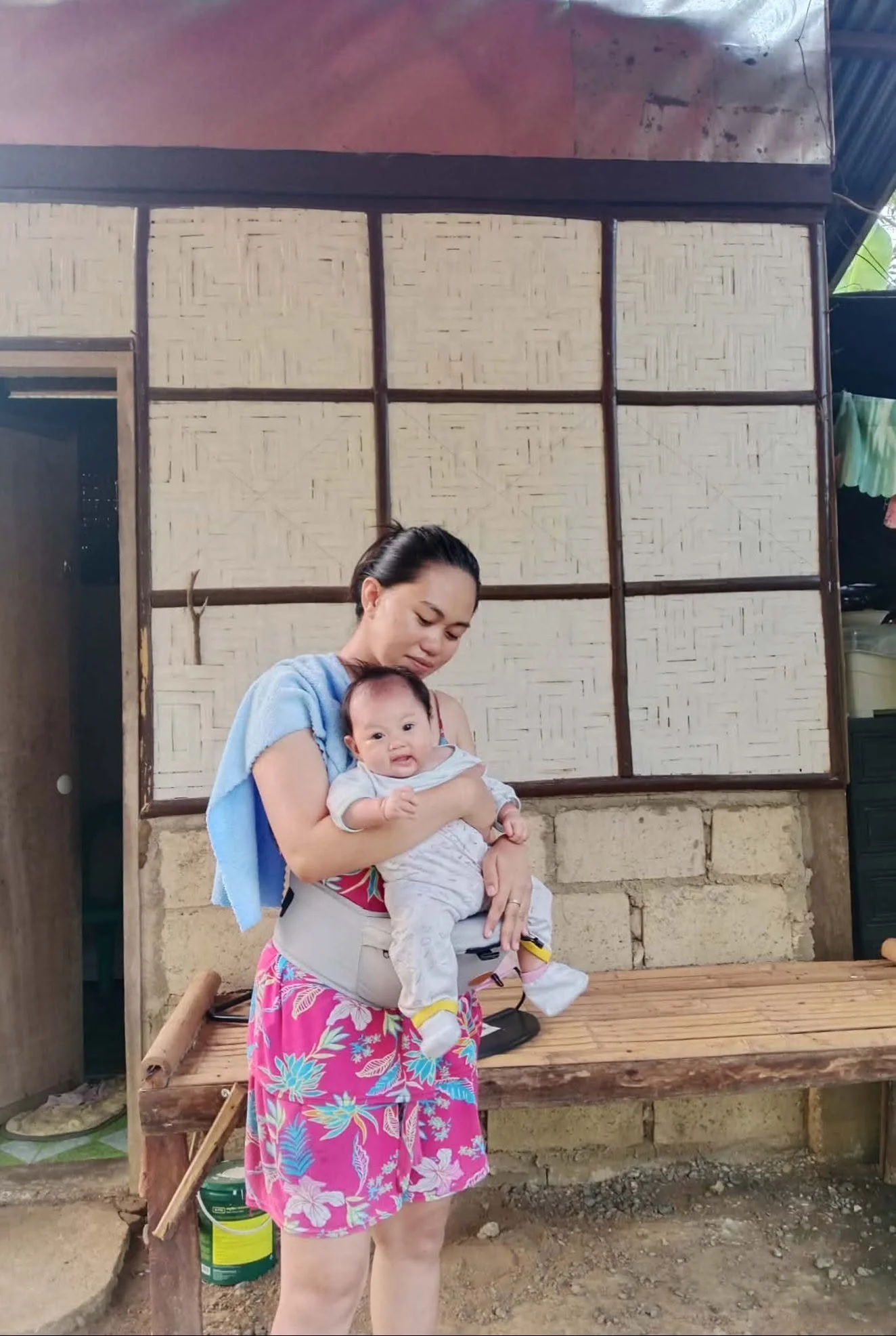 A woman with dark hair in a ponytail holding a smiling baby. The woman is wearing a pink floral skirt and a gray top. The baby is dressed in a light outfit and yellow shoes. They are standing in front of a wooden bench with a building made of brick a