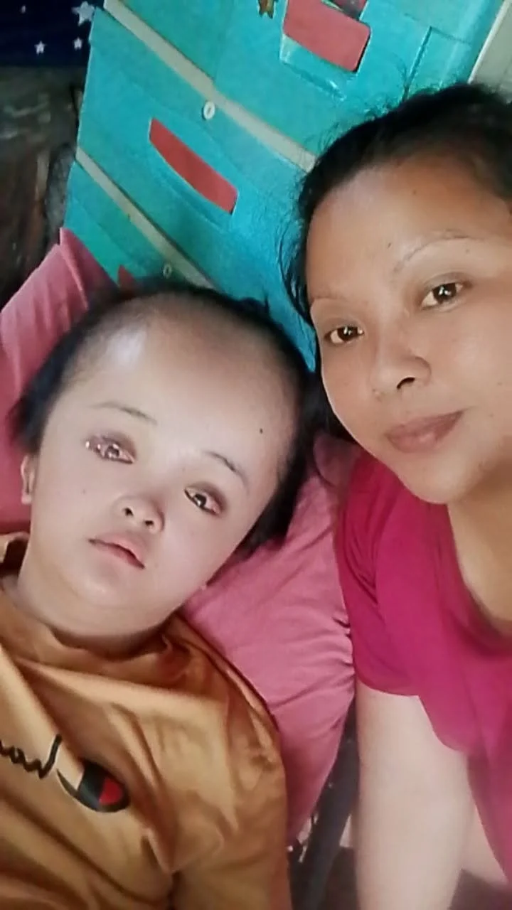 Two women lying close together, taking a selfie. One woman has short hair and is wearing a pink shirt, the other has a shaved head and is wearing a brown shirt. They are indoors, with a colorful object or furniture in the background.