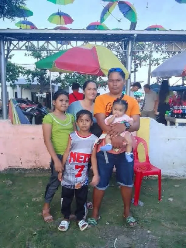 A family of five standing outdoors under colorful umbrellas at a park or fair. The family includes two women, one man, and three children. The man is holding a young girl in his arms. The group is smiling, and the background features additional peopl
