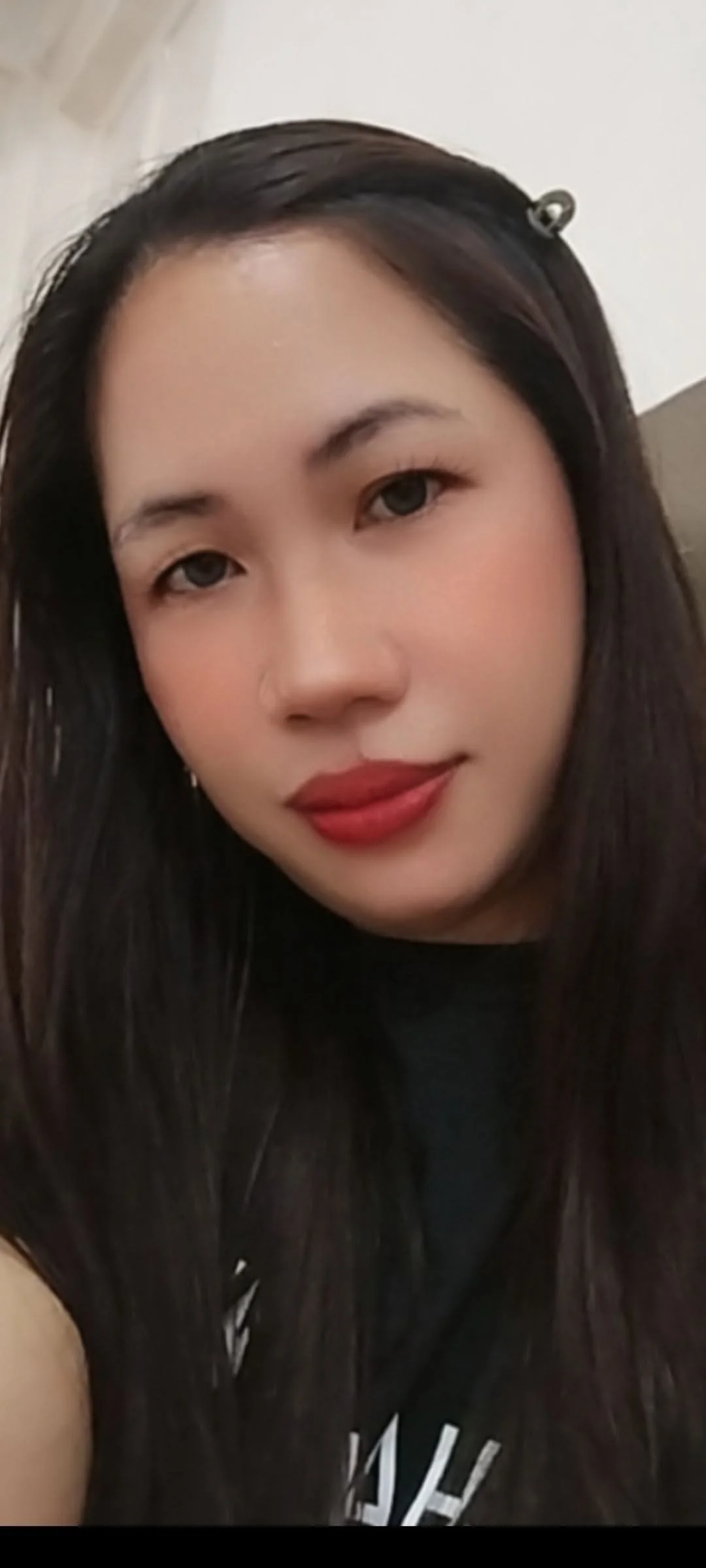 A close-up of a young woman with long black hair, light skin, red lipstick, and minimal makeup, smiling slightly.