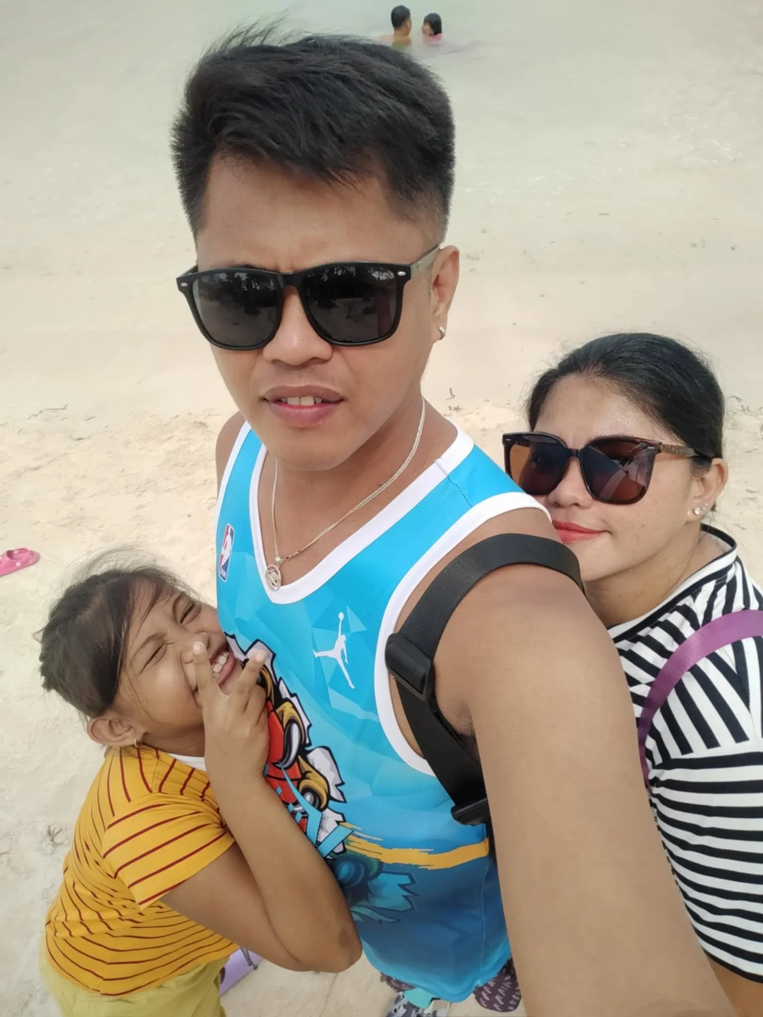 A group of people at the beach with a man and woman in the foreground overlapping, and children partially visible. The man is wearing sunglasses, a sports jersey, and a necklace. The woman is wearing large sunglasses and a striped shirt. A child in a
