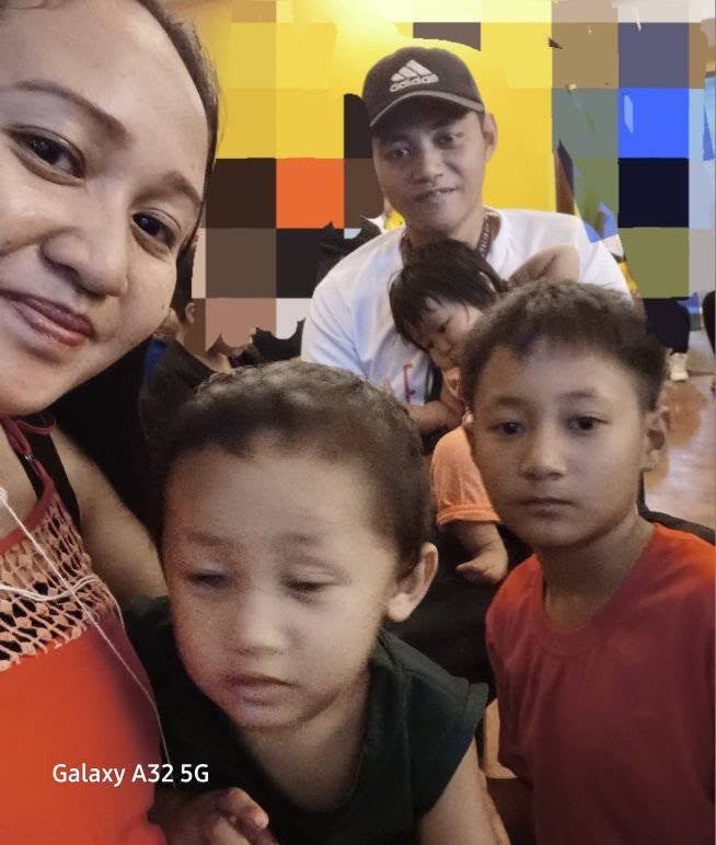 A group of five people, including two women and three children, in an indoor setting with brightly colored walls. The woman on the left is smiling, and the others have neutral or serious expressions. One child in a red shirt is prominently in the for