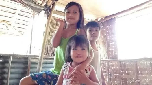Three children inside a rustic wooden structure, with sunlight streaming through gaps in the walls. The oldest girl has long dark hair and wears a green top, the middle girl, who appears younger, has short dark hair and wears a light-colored shirt, a