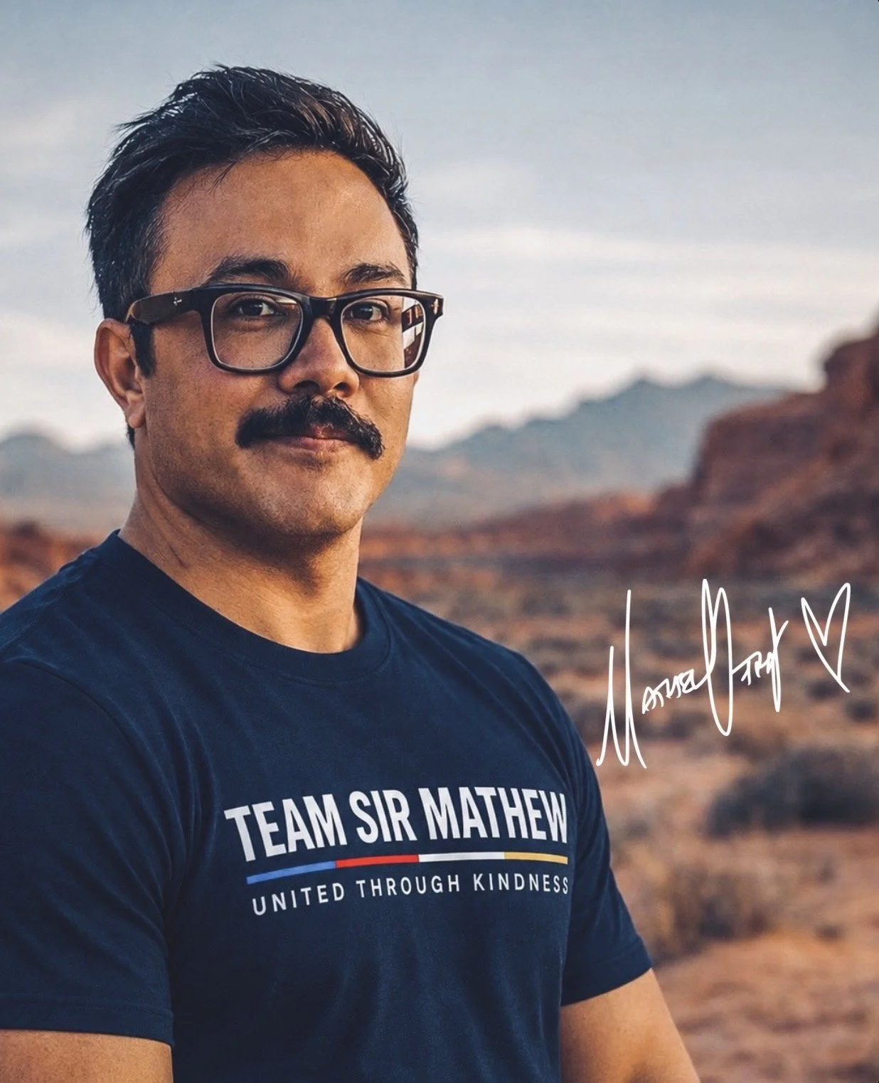 A man with glasses and a mustache standing outdoors in a desert landscape, wearing a navy blue t-shirt with 'Team Sir Mathew' and a signature with a heart on it.