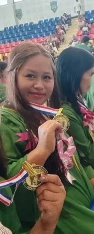 Young girl smiling and holding a gold medal at a sports event, seated indoors with other children and spectators in the background.