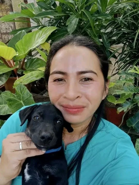 A woman with dark hair wearing a blue shirt holding a black puppy with a small white patch on its chest, surrounded by green plants.