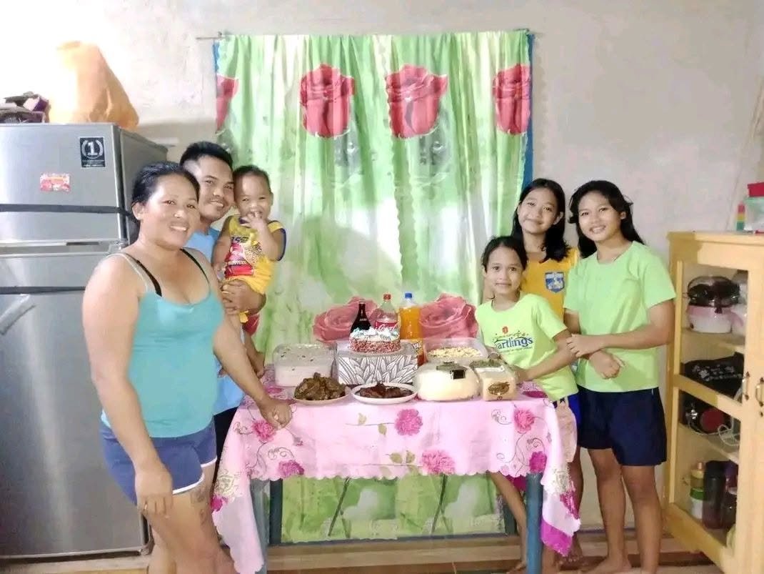 A group of seven people, including three adults and four children, standing around a table with food in a kitchen. The scene features a refrigerator, a green curtain with red flowers as a backdrop, and a wooden shelf on the right. They appear to be c
