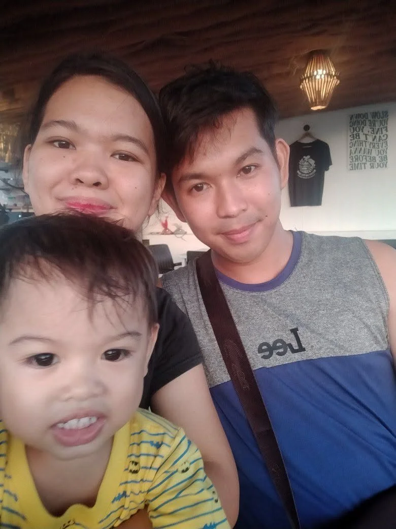 A group selfie of a woman, a young man, and a toddler inside a restaurant or cafe. The woman is smiling, the man has a slight smile, and the toddler is making a face. The background has a wooden ceiling, hanging light fixture, and a t-shirt hanging o
