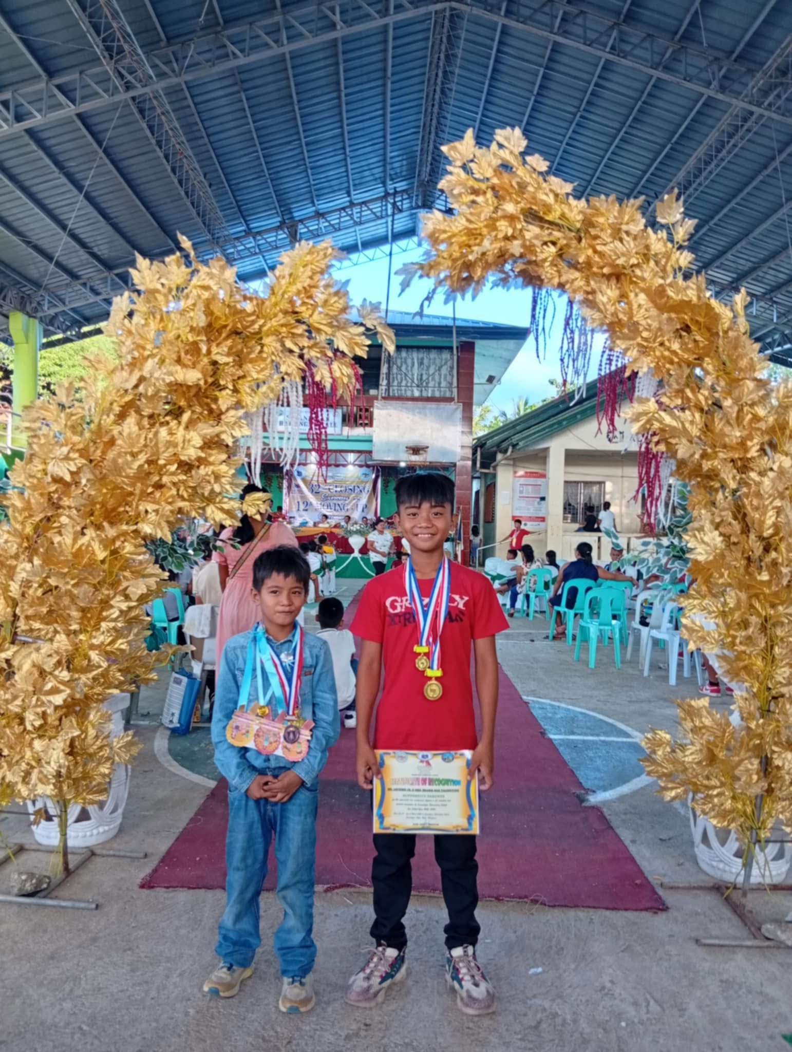 Two boys standing under a decorative arch made of gold-colored leaves at an outdoor event, with one boy wearing medals and holding a certificate, and the other boy smiling, all in front of a tent and chairs with other people and decorations in the ba