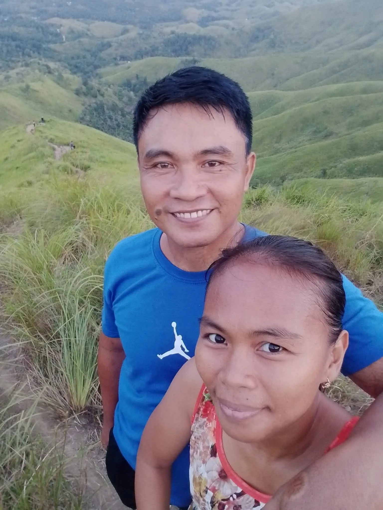 A smiling man and woman taking a selfie outdoors in a lush, green hilly landscape.