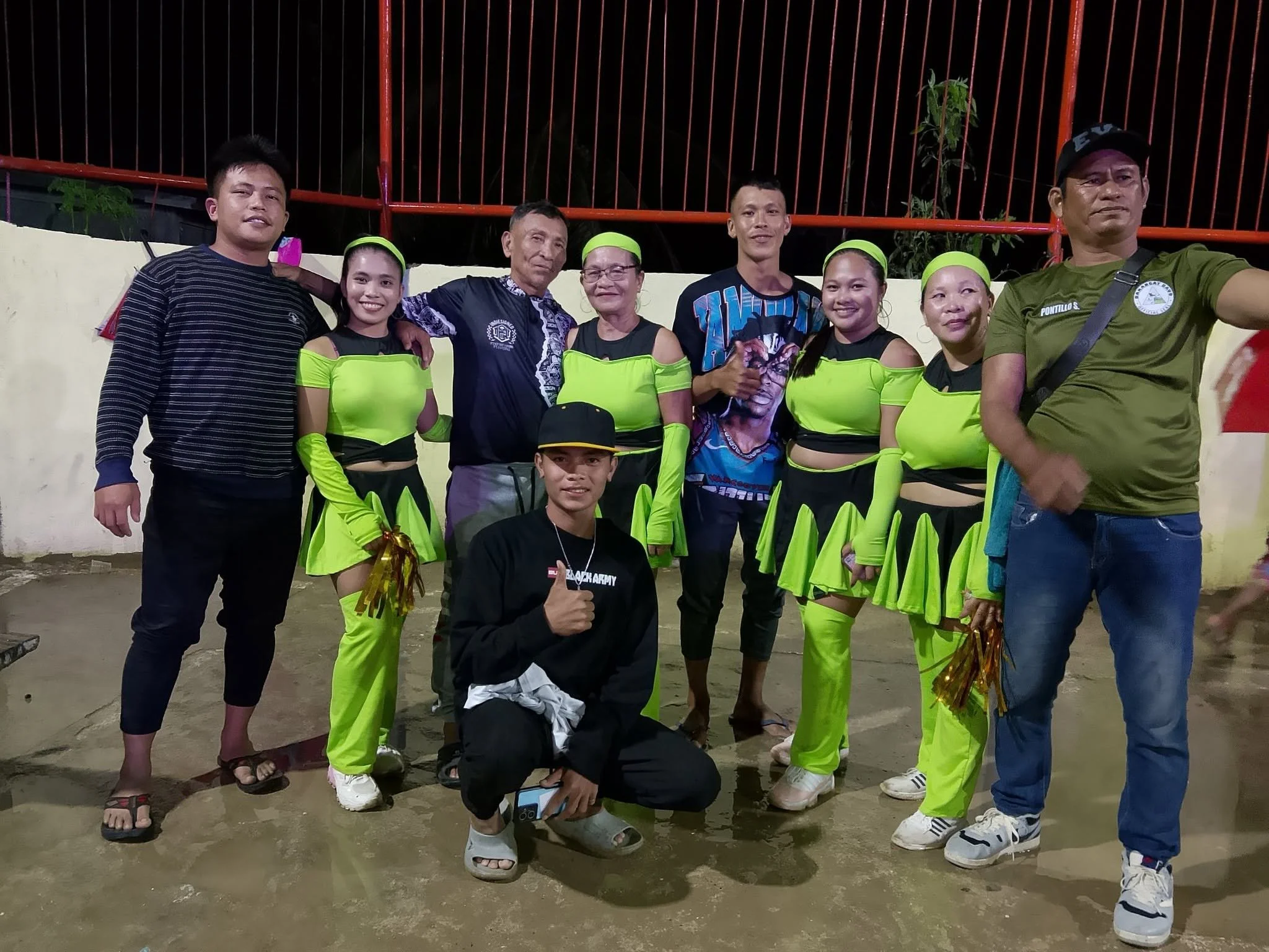Group of people, including cheerleaders or performers in bright green costumes, showing thumbs up and smiles, gathered outdoors at night with a orange fence and plants in the background.