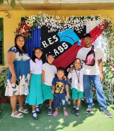 Group of five children and one adult standing in front of a graduation-themed backdrop with a blue cap and red ribbon, celebrating graduation at a school event.