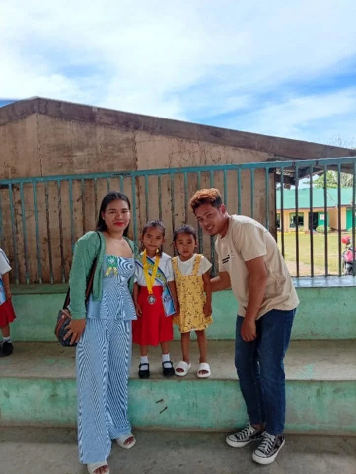 Two women and two young girls standing outside in front of bamboo fence; one girl wears a medal around her neck; they are smiling.